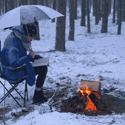 Ihminen istuu retkituolilla lumisateessa sateenvarjon alla ja lukee.