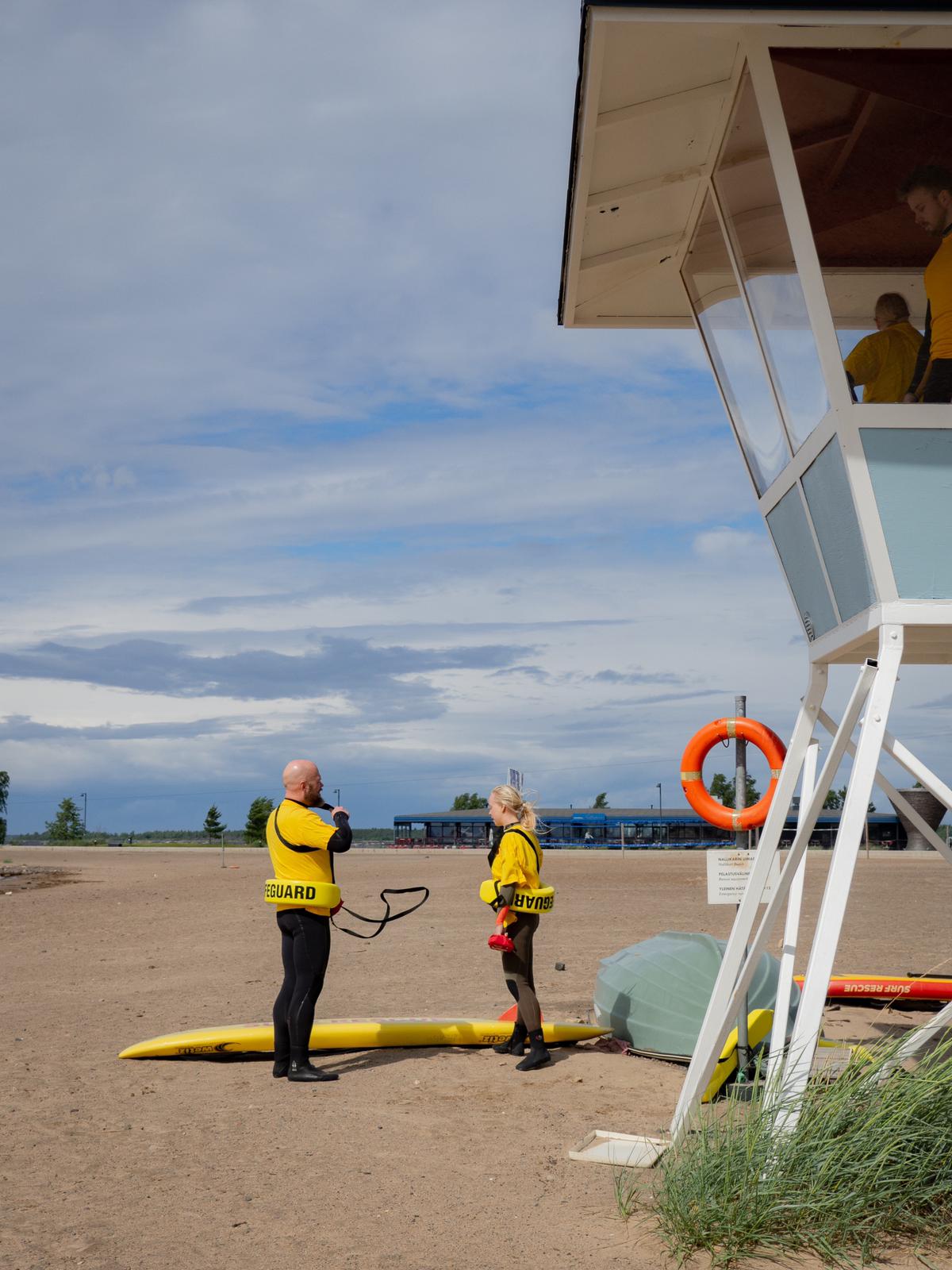 Kaksi keltapaitaista rantavalvojaa seisovat rannalla valvontatornin vieressä. Valvontatornissa on myös rantavalvojia.